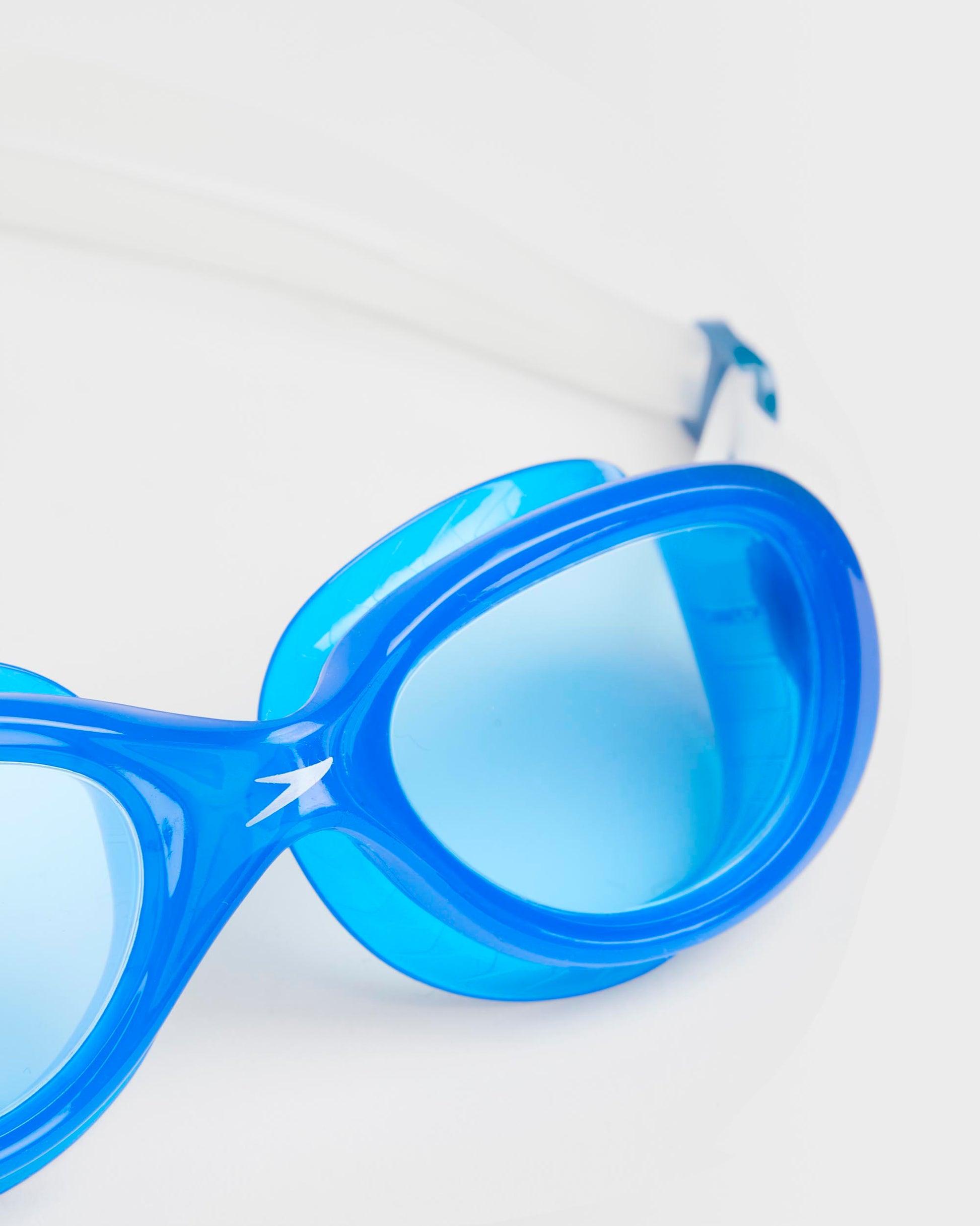 Close up of a pair of blue swimming goggles with a white strap, viewed from the front on a white background.